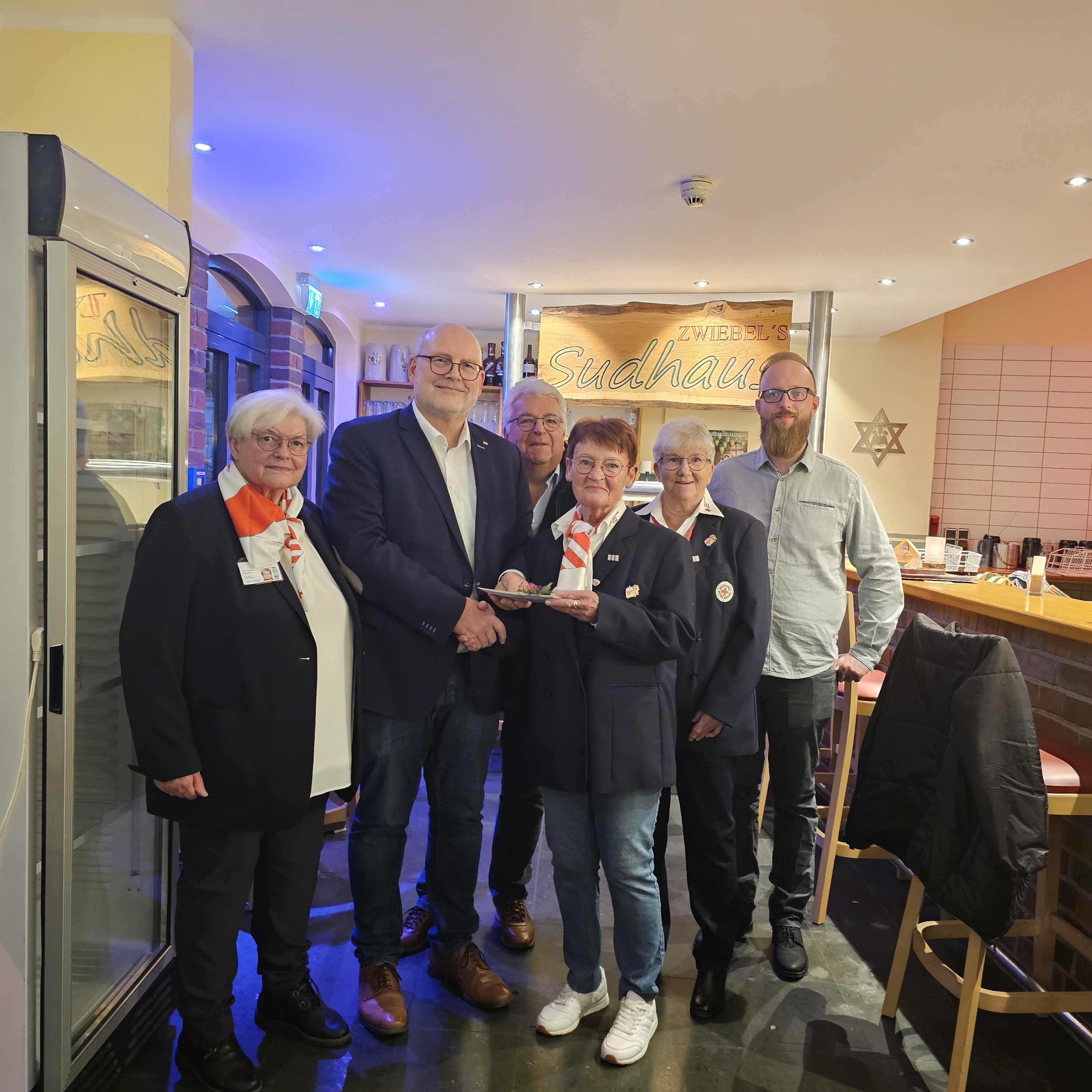 Sechs Personen stehen in einem Restaurant. Eine Frau bekommt einen Blumenstrauß überreicht.