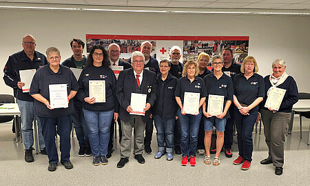 MItgliederversammlung 15 Personen stehen mit Ihren Urkunden in einer zweierreihe in einem beleuchteten Raum beim DRK Soest.